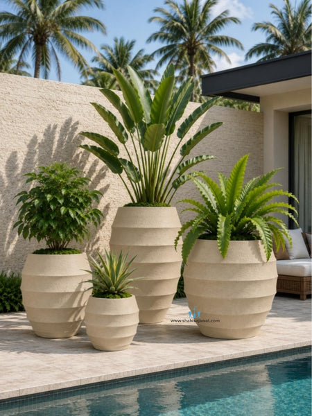 Three large beige ribbed planter pots with green plants arranged on a poolside patio in front of a stucco wall and palm trees.